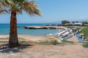 A beach with a palm tree, a rocky shoreline, and a row of beach umbrellas and lounge chairs. - Cretan Seaside Boutique Hotel (Hersonissos)