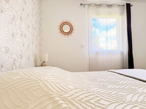 Un dormitorio con pared estampada, un adorno mural con forma de sol, una cama con manta texturizada, una lámpara de noche y una ventana con cortinas.