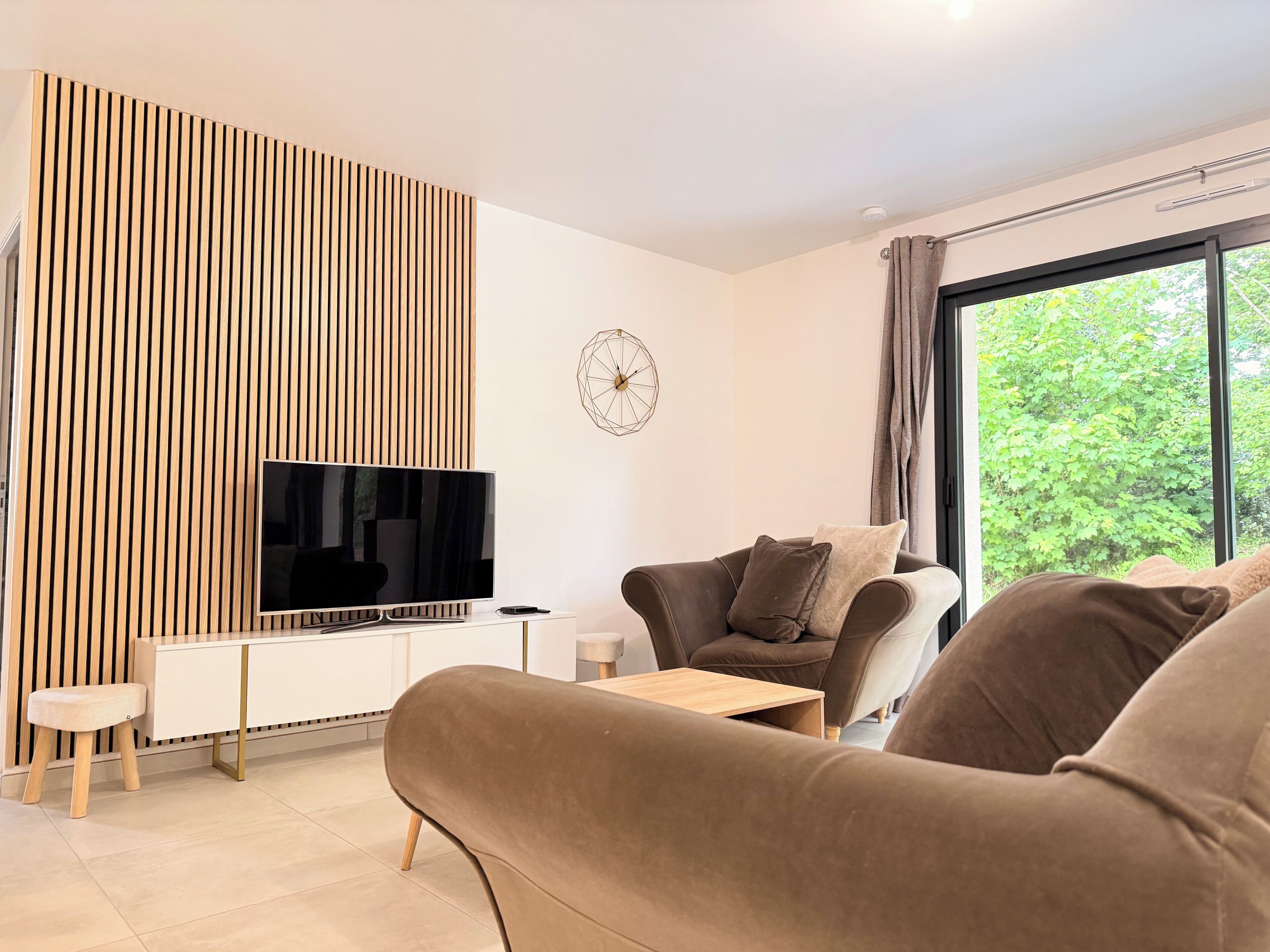 Ein modernes Wohnzimmer mit Flachbildfernseher, einer Couch, einem Couchtisch und einem großen Fenster mit Blick auf Grünflächen.