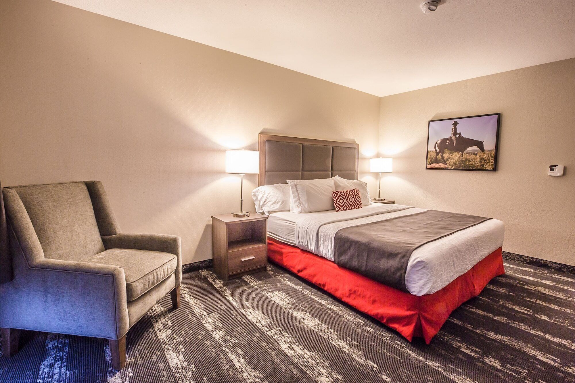 A hotel room with a bed, two bedside lamps, a chair, a nightstand, a carpet, and a wall-mounted picture.