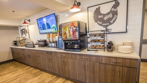 A kitchen area with a coffee machine, juice dispensers, a TV displaying a news channel, and a wall-mounted chicken silhouette. - Ramada Cochrane (Cochrane)