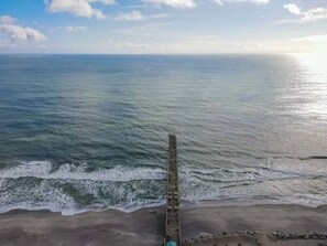 Blick auf die Küste mit einem hölzernen Steg, der ins Meer führt, einem Sandstrand und einem klaren Himmel.