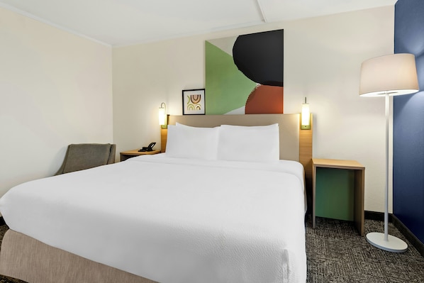 A hotel room with a large bed, a desk, a chair, a lamp, and a wall with a colorful abstract painting.