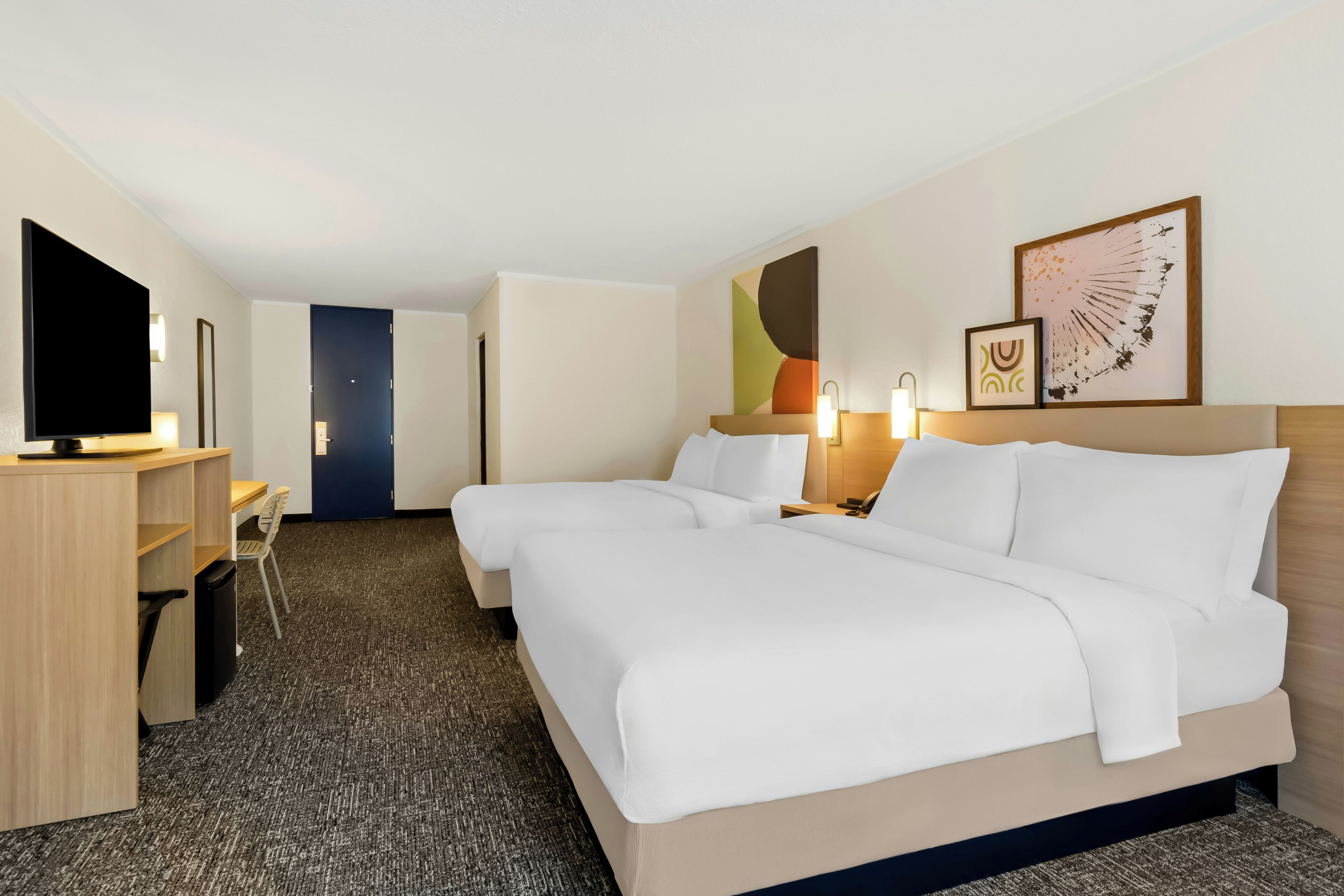 Quarto de hotel com duas camas, uma escrivaninha, televisão e obras de arte nas paredes.