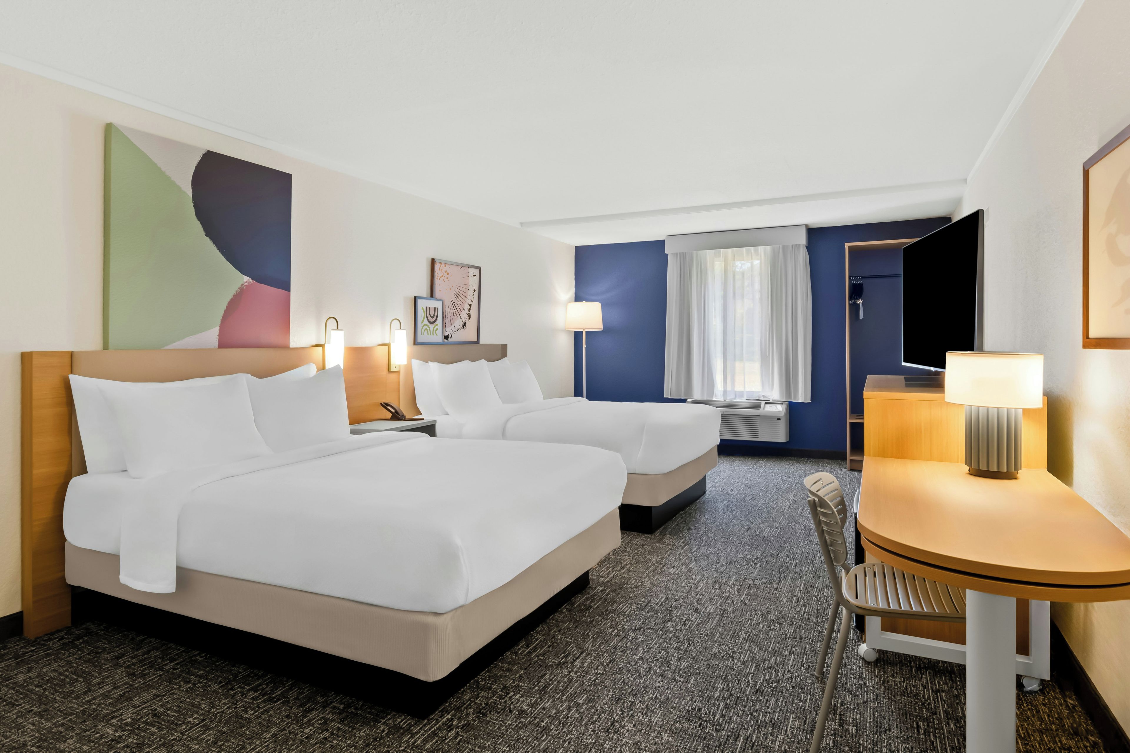 Habitación de hotel con dos camas, un escritorio, una silla, un ventanal grande con cortinas y obras de arte en las paredes.