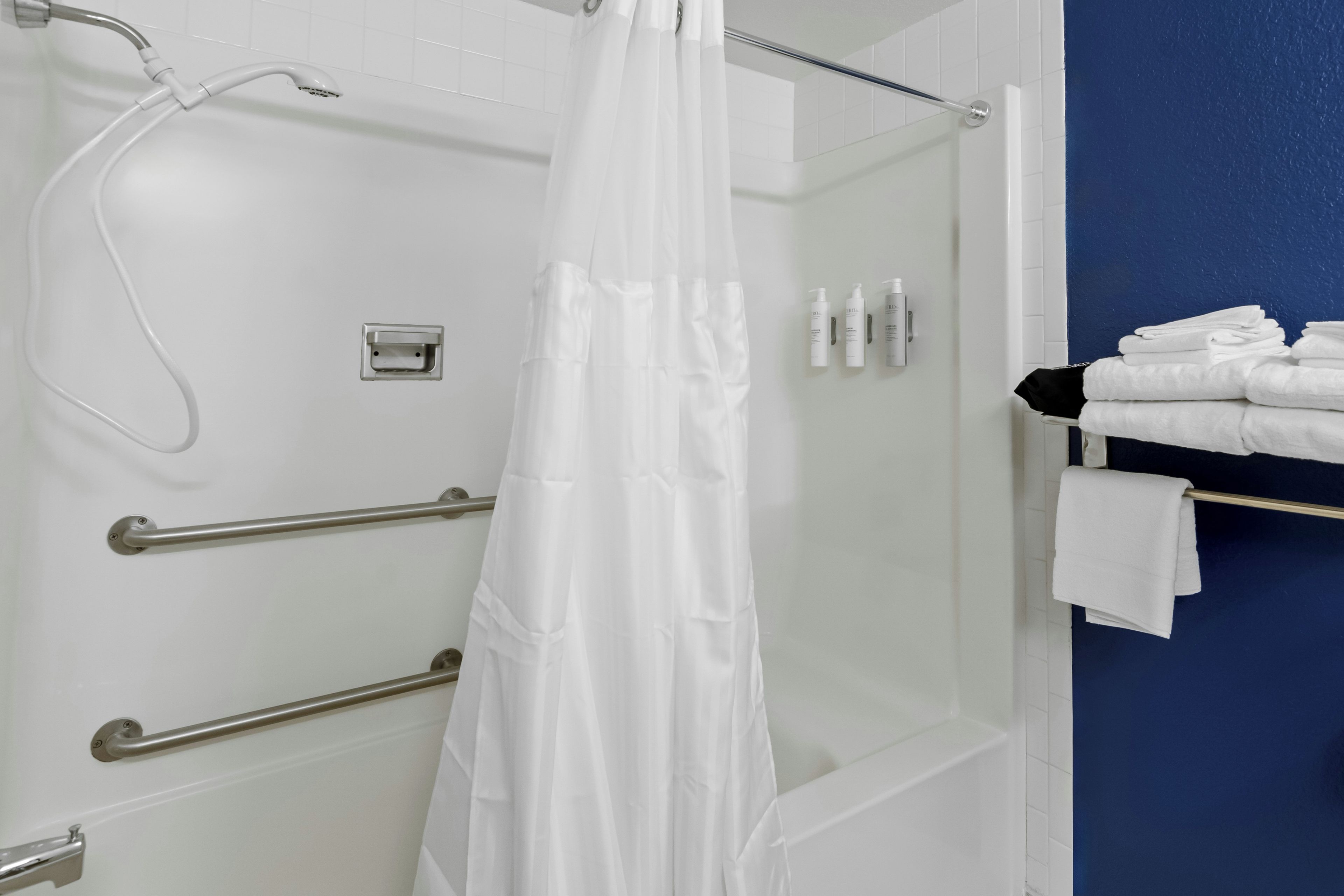 A shower area with a white shower curtain, grab bars, and a towel rack with folded towels.