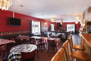 A restaurant interior with red walls, multiple tables with checkered tablecloths, wooden chairs, and a bar area.