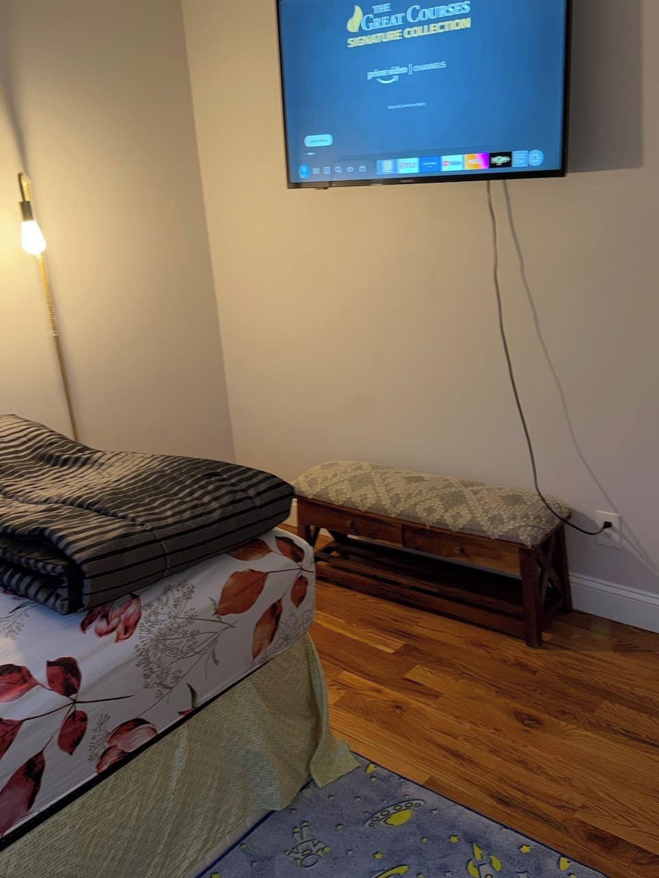A room with a bed, a bench, a wall-mounted television displaying a course collection, and a wooden floor.