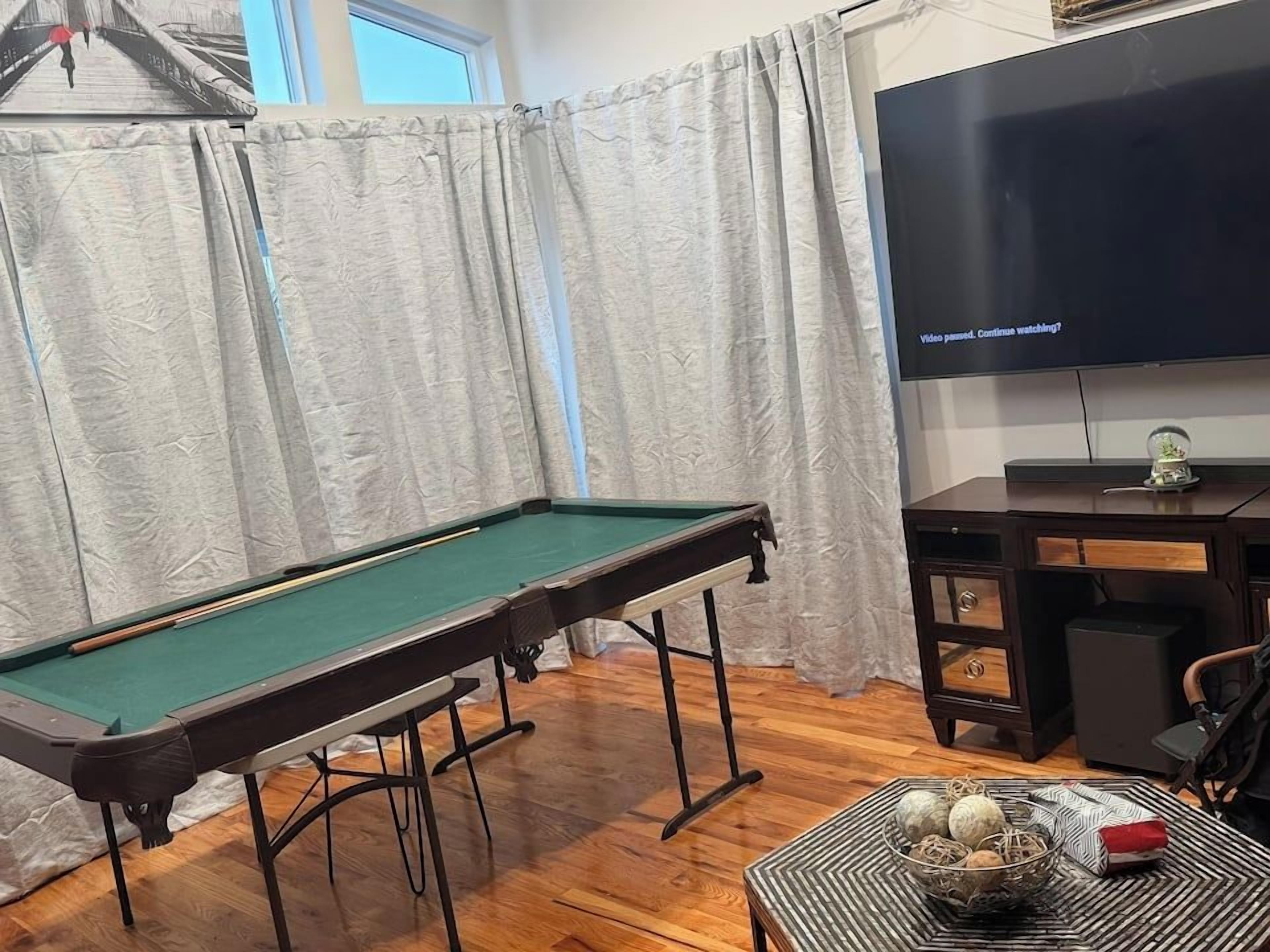 A pool table with a green felt surface, a television mounted on the wall, and a small table with decorative items.
