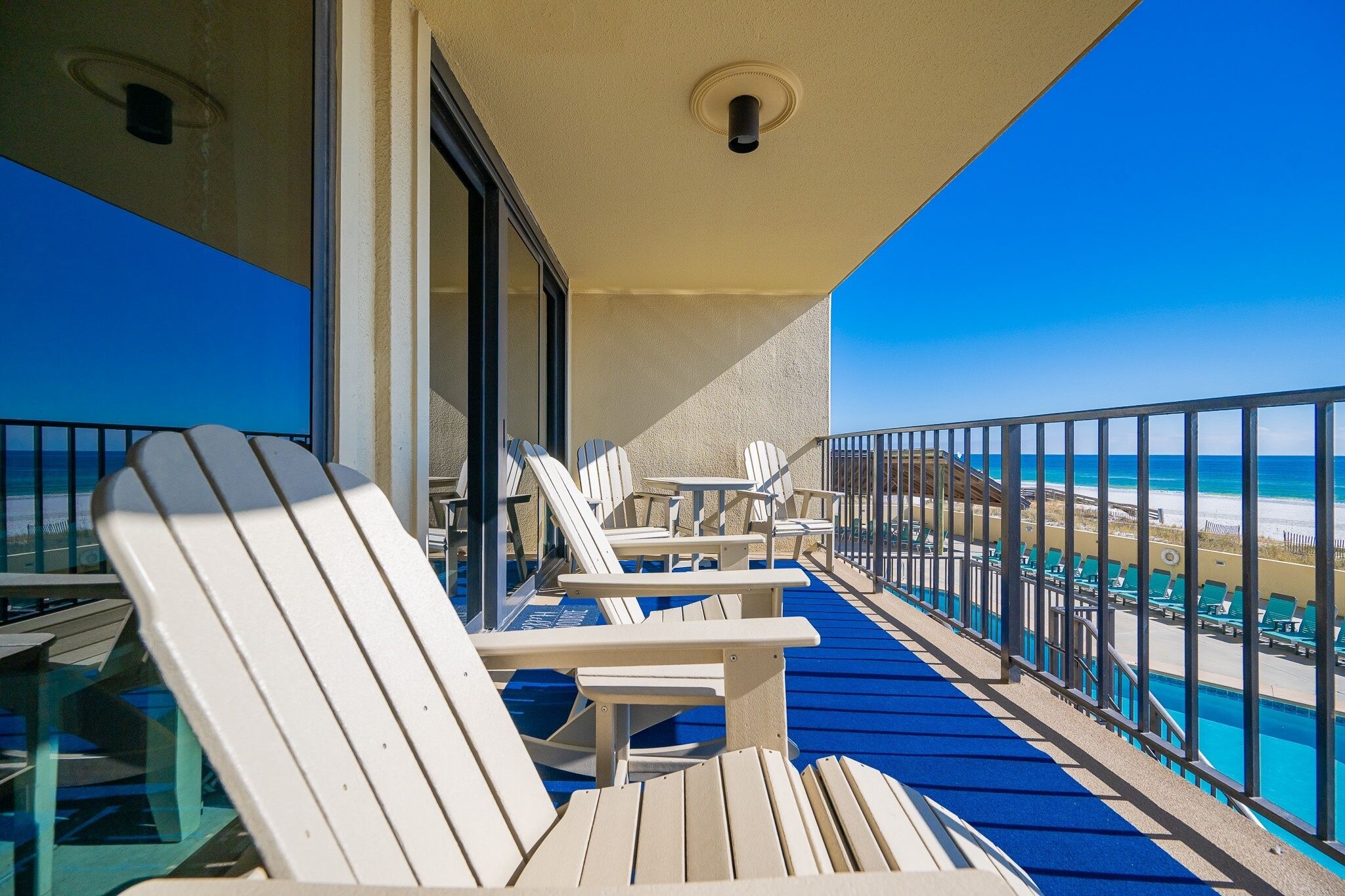 A balcony with white Adirondack chairs, a blue carpet, and a view of the beach.