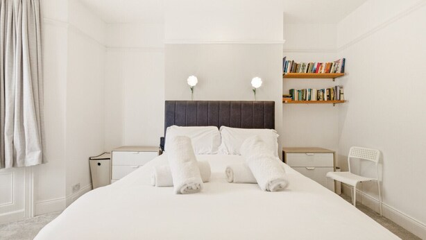 Una cama bien hecha con sábanas blancas y toallas dobladas, una mesita de noche con lámpara, una estantería con libros y una silla blanca.