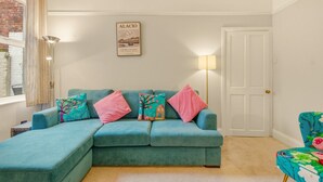Un sofá color turquesa con cojines rosa y estampados florales, un cuadro en la pared, una lámpara y una puerta.