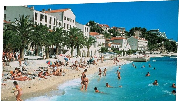 A beach with people swimming and sunbathing, palm trees, and buildings in the background. - Seafront Studio Apartment with Balcony (Podgora)