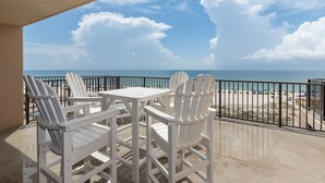 Ein Balkon mit weißen Adirondack-Stühlen und einem Tisch, mit Blick auf einen Strand unter blauem Himmel, durch eine Ziegelmauer vom Balkon getrennt.