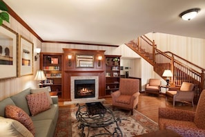 A living room with a fireplace, bookshelves, a staircase, and patterned furniture. - Country Inn & Suites by Radisson, Manchester Airport, NH (Bedford)