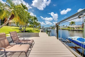 A deck with chairs, a boat docked by the water, and palm trees in the background. - Villa Tip-Toe Inn - charming 3 BR/ 2 bath direct access waterfront home w. heated pool, paddle boat, foosball table, dart board, fishing poles, and bikes (Cape Coral)