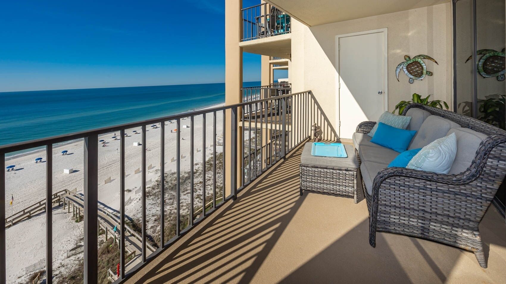 Un balcon avec vue sur la plage, un canapé en osier agrémenté de coussins et une porte décorée d’une représentation de tortue.