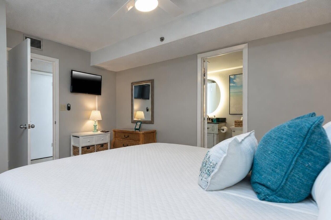 A hotel room with a large bed, a television, a bedside table with a lamp, a dresser, and a mirror.