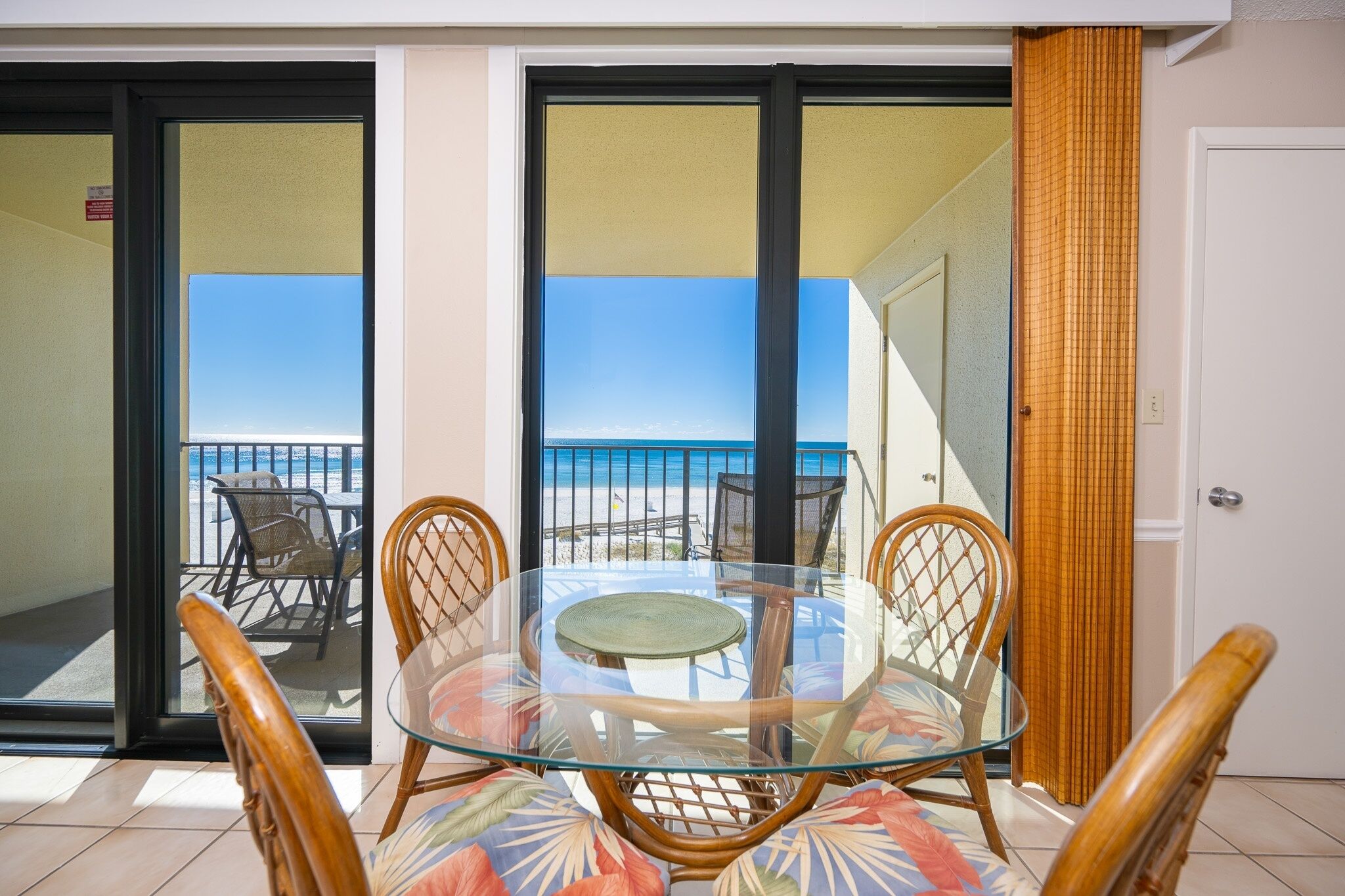 Een tafel met glazen blad en rieten stoelen, een schuifpui naar een balkon met uitzicht op het strand.