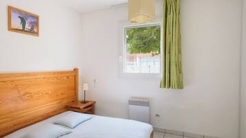 Un dormitorio con cabecera de madera, cama con ropa de cama blanca, mesita de noche con lámpara, ventana con cortinas y radiador.