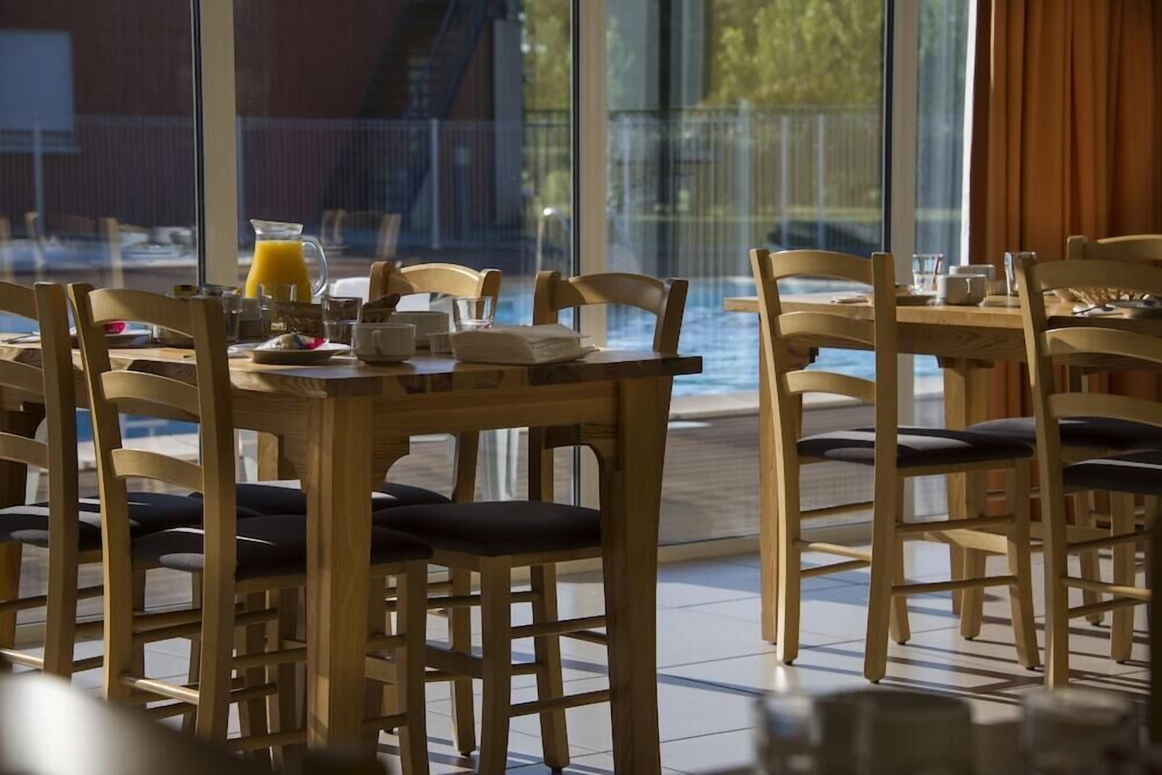 A breakfast setting with wooden tables and chairs, a pitcher of orange juice, and a view of a swimming pool through large windows.