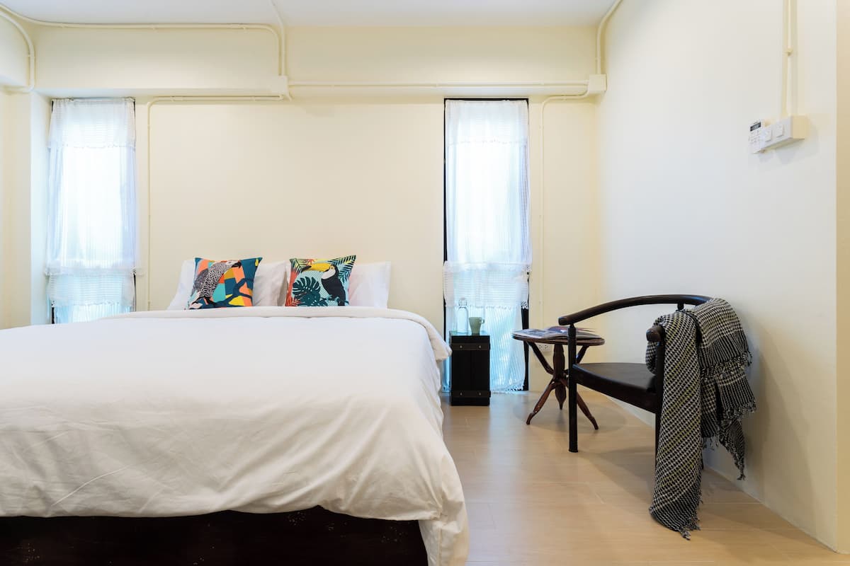 Un dormitorio con cama, dos ventanas con cortinas, una silla y una mesita.