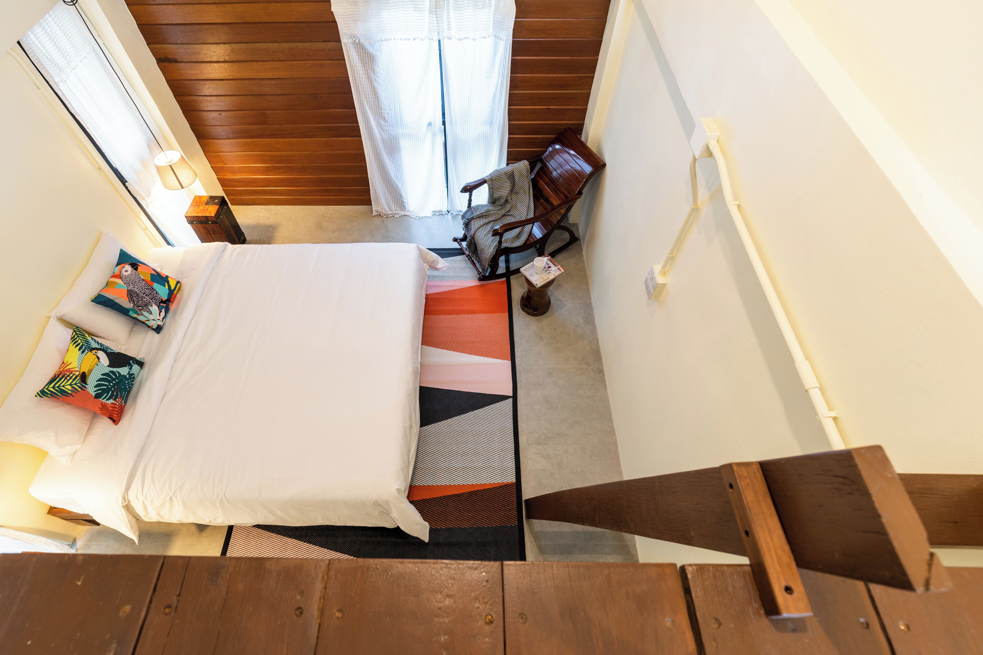 Een slaapkamer met een bed, kussens, een stoel, een klein tafeltje en een raam met gordijnen.