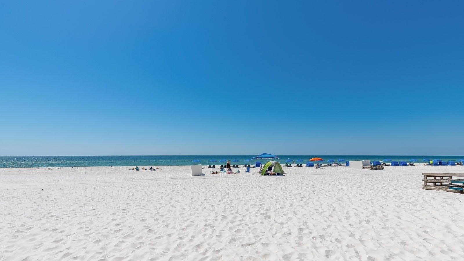 A wide sandy beach with a clear blue sky, several beach umbrellas, and a few tents.