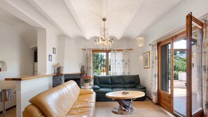 A living room with a sofa, a table, a chandelier, and a view through a glass door. - House with Spacious Terrace and Private Barbecue (Begur)