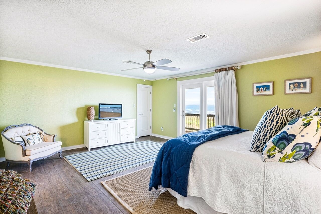 A bedroom with a bed, a ceiling fan, a television, a dresser, and a view of the outdoors.