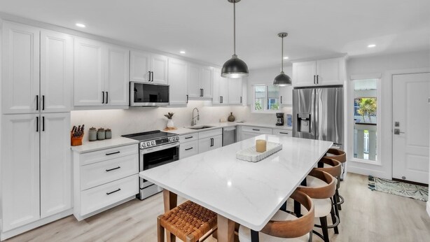 Una cocina moderna con muebles blancos, electrodomésticos de acero inoxidable, una isla central con asientos, iluminación colgante y vistas al exterior.