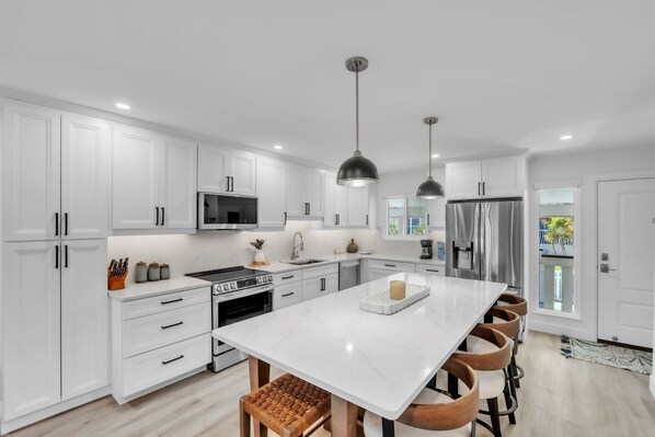 A modern kitchen with white cabinetry, stainless steel appliances, a central island with seating, pendant lighting, and a view to the outside. - Runaway Bay 276 2 Bedroom Home (Bradenton Beach)