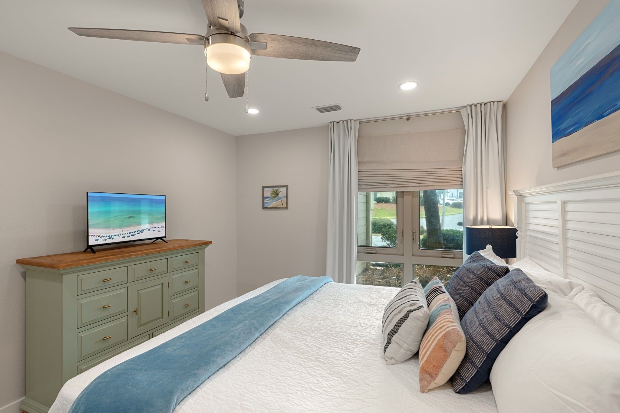 Une chambre à coucher avec un lit, un téléviseur posé sur une commode, un ventilateur de plafond et une fenêtre avec des rideaux.
