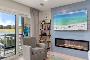 Ein Wohnzimmer mit Kamin, großem Fernseher, Bücherregal und Blick auf einen Strand durch eine Glasschiebetür.