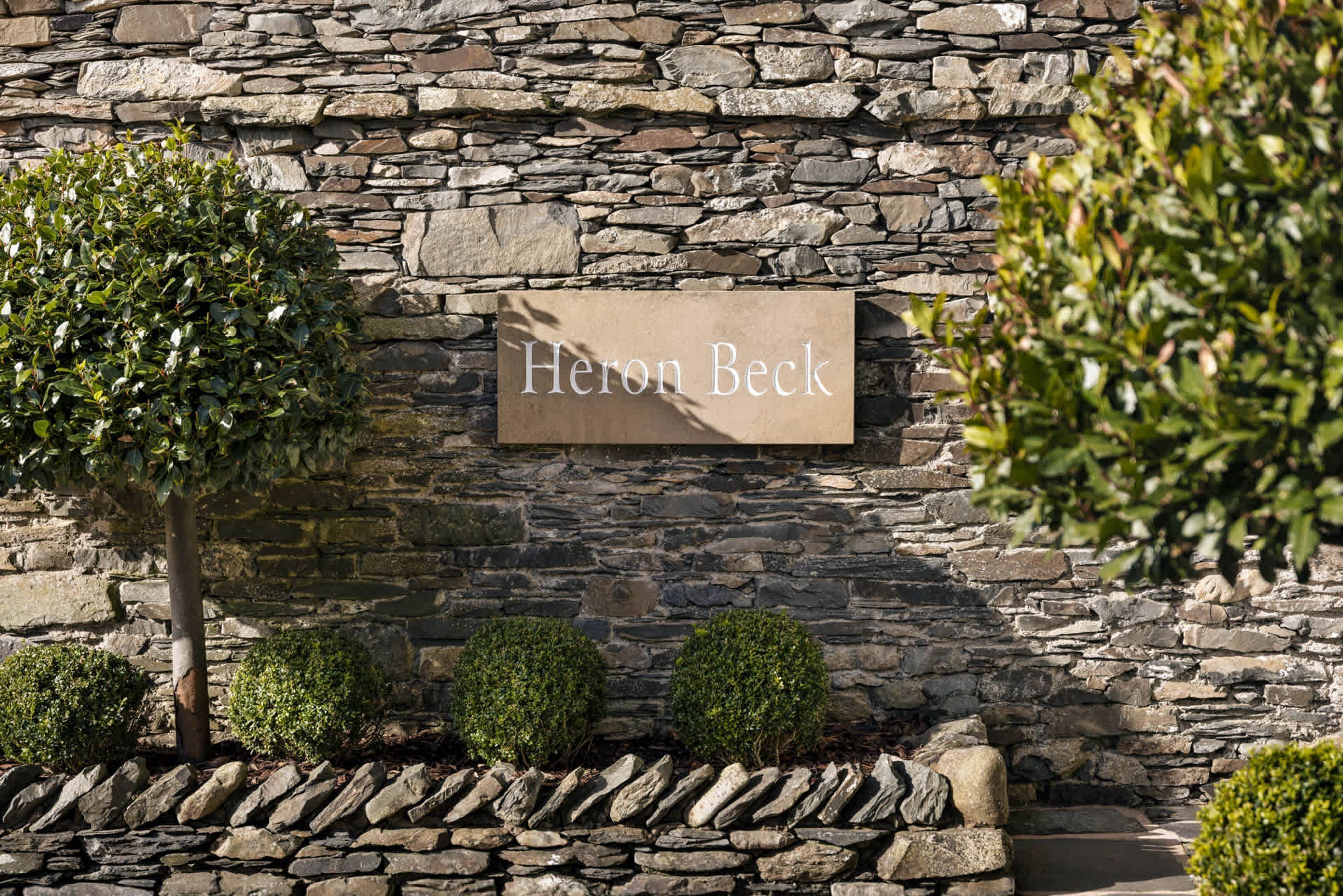 A stone wall with a sign reading 'Heron Beck' and a small tree in front.