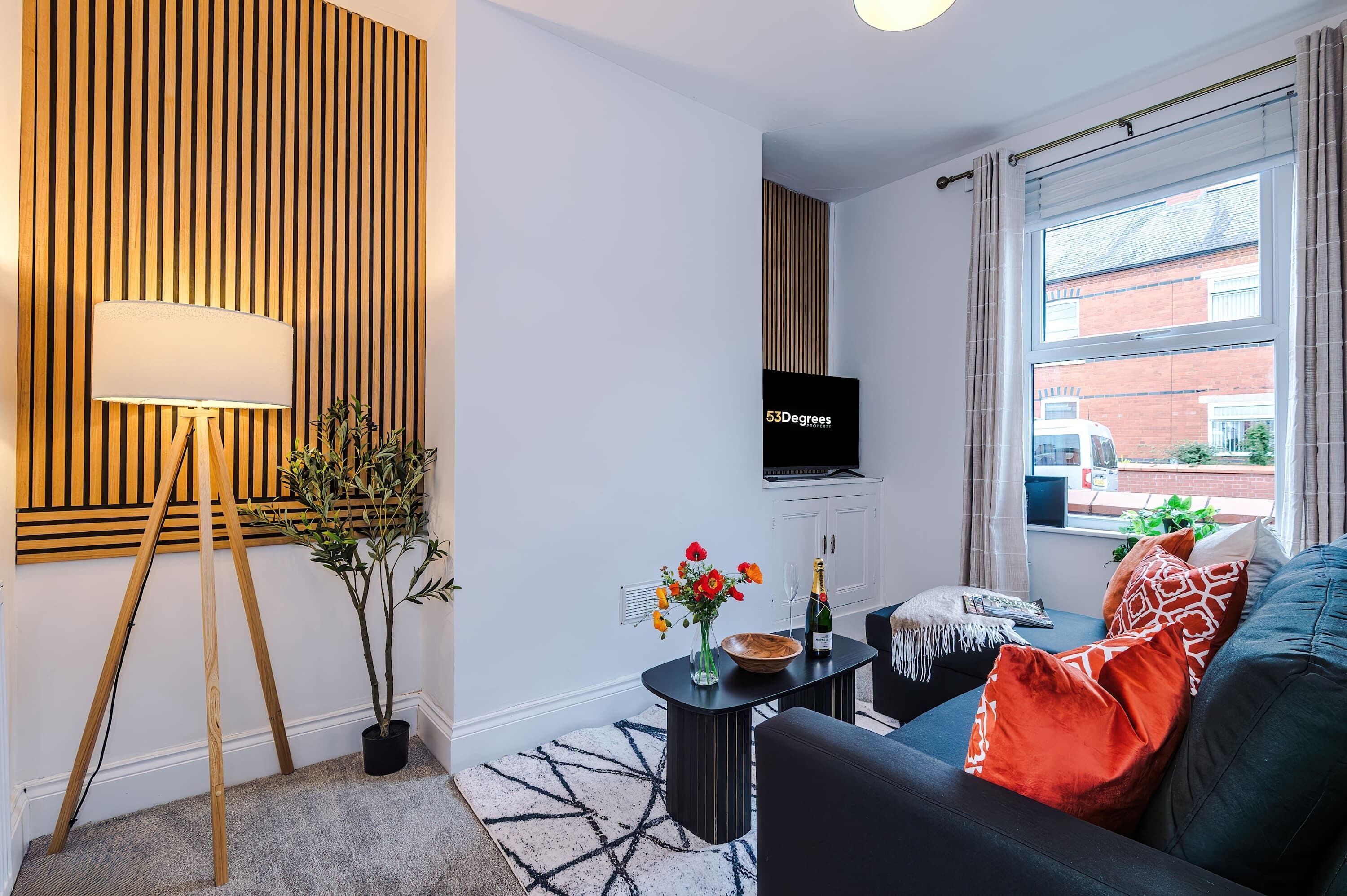 A modern, inviting living room with a sleek dark sofa, bold orange and patterned cushions, and a TV positioned for easy viewing. Fresh flowers and champagne on the table add a touch of luxury, while framed botanical prints bring a natural feel.