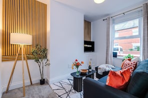A modern living room with a TV mounted on the wall, a potted plant, a small table with a vase of flowers, and a sofa with cushions.