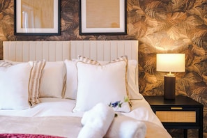 A bed with white linens and striped pillows, a bedside lamp, and a wall with a leaf-patterned wallpaper.