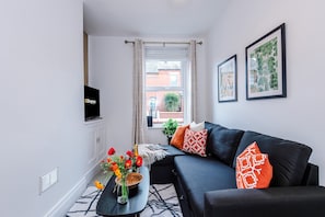 A modern living room with a black sofa, a TV mounted on the wall, a window with curtains, and framed artwork on the wall.