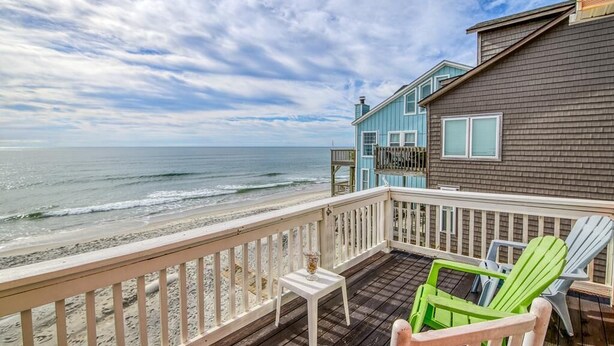 Une terrasse en bois avec une rambarde donnant sur une plage, une petite table et des fauteuils Adirondack aux couleurs vives.