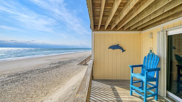 A wooden deck with a blue chair, a dolphin silhouette on the wall, and a beach view. - Oceanfront Escape at Surf Condo 814 – Relaxation Starts Here (Surf City)