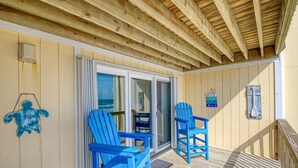 A wooden deck with two blue chairs and a table, a glass door leading to a shaded area, and a wooden ceiling. - Oceanfront Escape at Surf Condo 814 – Relaxation Starts Here (Surf City)