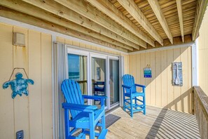 A wooden deck with two blue chairs and a table, a glass door leading to a shaded area, and a wooden ceiling. - Oceanfront Escape at Surf Condo 814 – Relaxation Starts Here (Surf City)