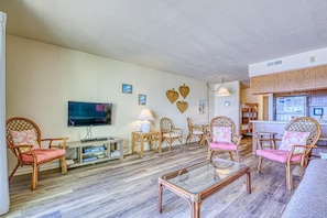 A living room with a TV, wooden chairs, a glass coffee table, and a kitchen area. - Oceanfront Condo – Surf City, NC (Surf City)