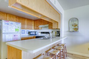A kitchen with wooden cabinets, a white countertop, a microwave, a stove, and a sink. - Oceanfront Condo – Surf City, NC (Surf City)