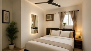 Et hotelværelse med seng, ventilator, spejl, et vindue med gardiner, et natbord med lampe og en potteplante.