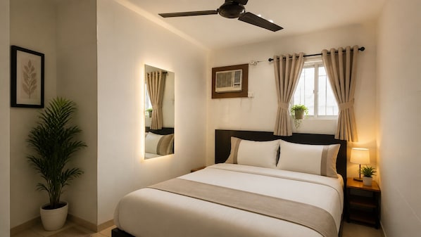 Quarto de hotel com cama, ventoinha no teto, espelho, janela com cortinas, mesa de cabeceira com abajur e planta em vaso.