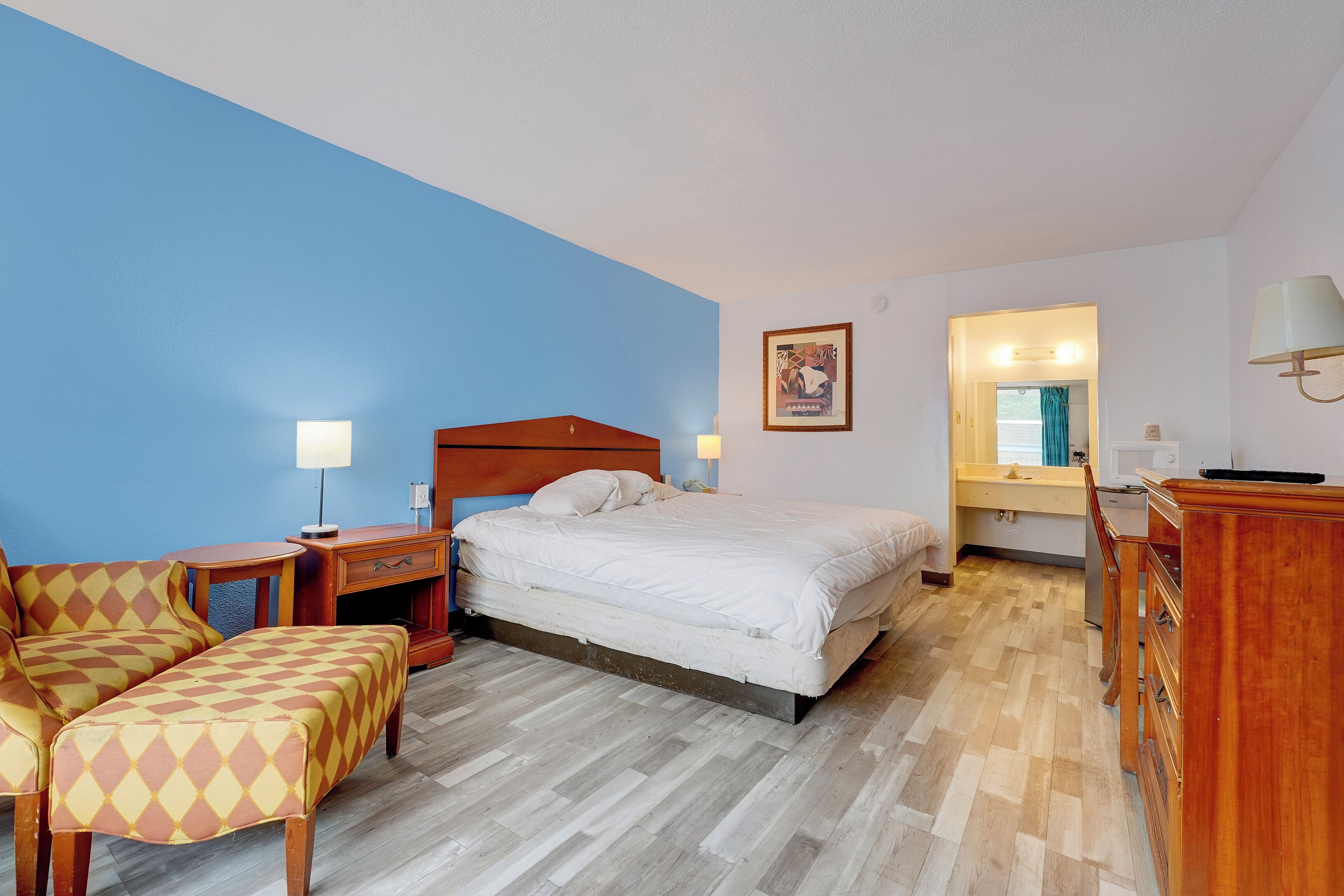 A hotel room with a bed, a desk, a chair, a bench, a dresser, a mirror, and a wall with a blue accent.