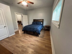 Ein Schlafzimmer mit Holzboden, einem Bett mit blauer Bettdecke, einem Deckenventilator und einem Fenster mit Jalousien.
