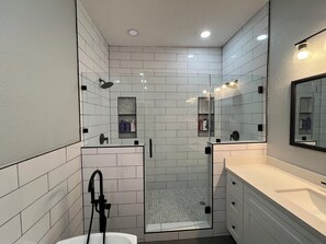 Un baño moderno con azulejos blancos, cabina de ducha de vidrio, tocador con espejo e iluminación empotrada en el techo.